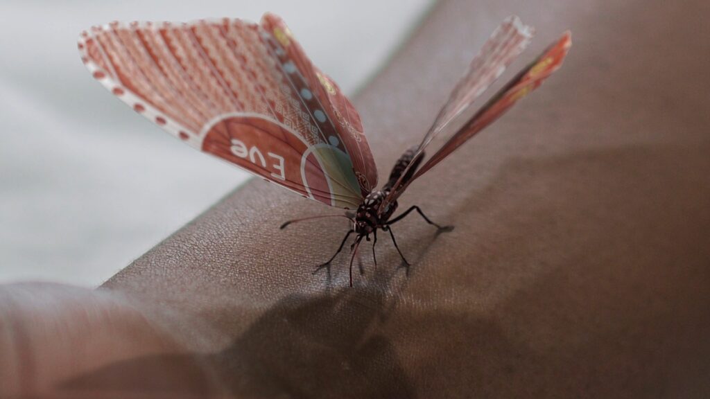 An extreme closeup of a genetically modified butterfly, red with white geometric designs on the wings, and the name Eve, sitting on the arm of a black person, veins show through the skin.