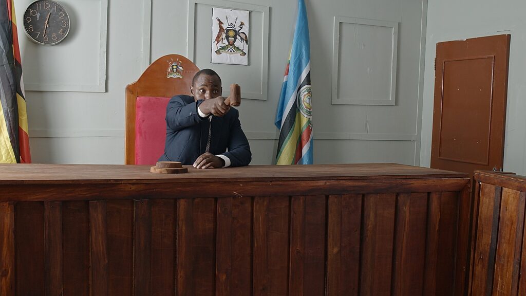 A magistrate in a film scene, holding his hammer as he points at somebody offscreen. It's a studio constructed set in a film studio in Kampala
