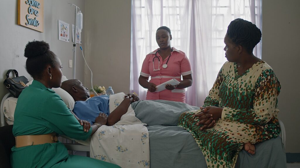 A hospital scene, a nurse stands above a patient, while two of the patient's relatives sit on the bed, looking on. It's a studio set built in a film studio in Kampala.