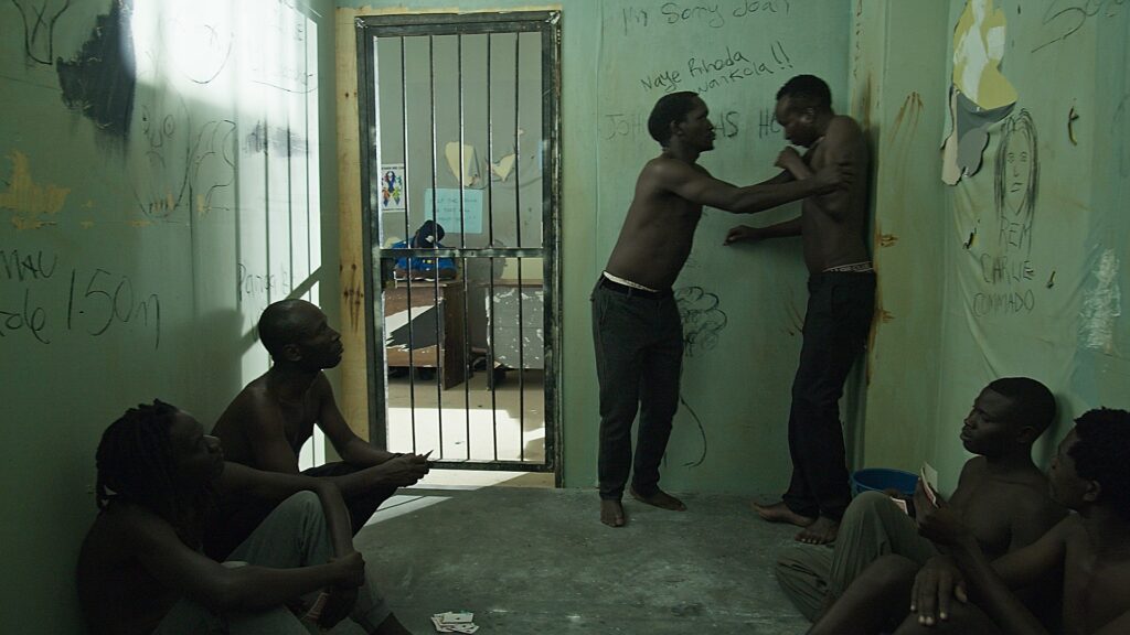 A jail scene, African men without shirts sit on the floor, while two are standing, one holds the other's neck. The cell is a studio set built in a film studio in Kampala.