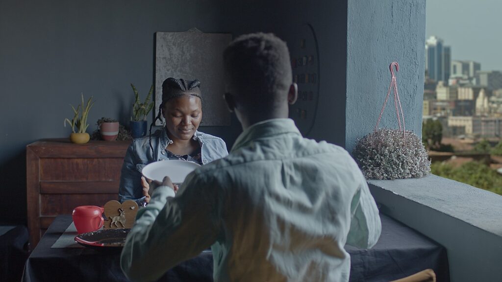 A restaurant scene, a couple sit with the man's back facing the camera. The woman is smiling. It's a scene constructed in a film set in a film studio in Kampala.